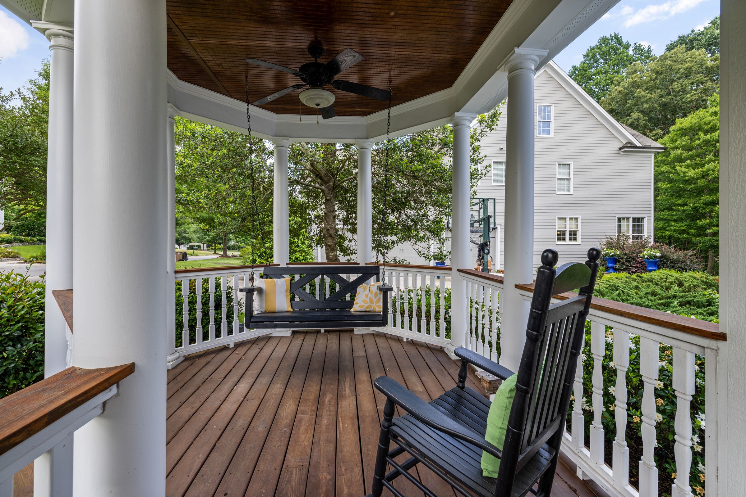 A wraparound porch in late-afternoon sun: a swing with yellow pillows, a rocking chair, and blue planters along the white rail.