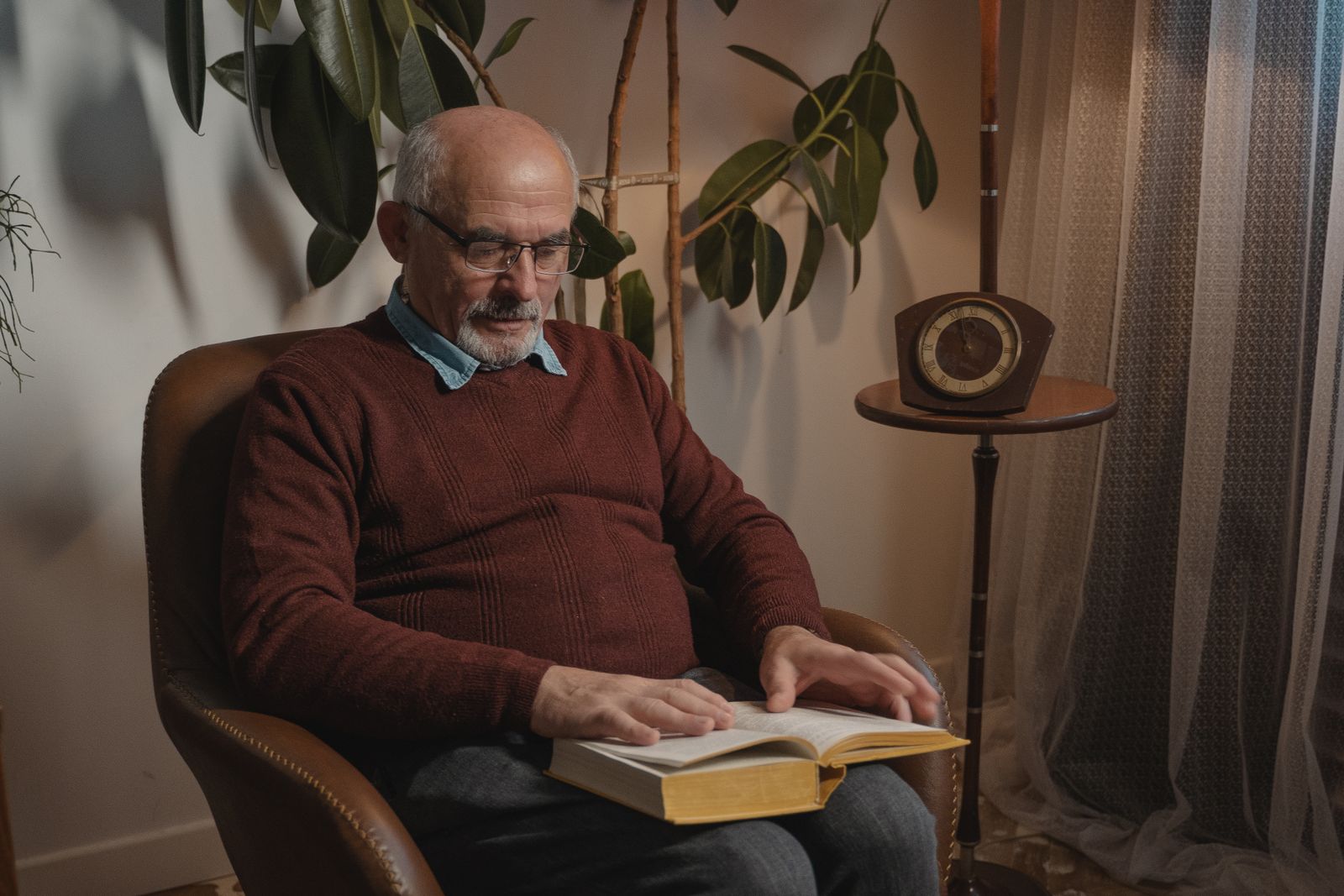 A man reading a hardback in a brown leather armchair, under warm lamplight beside a mantel clock.