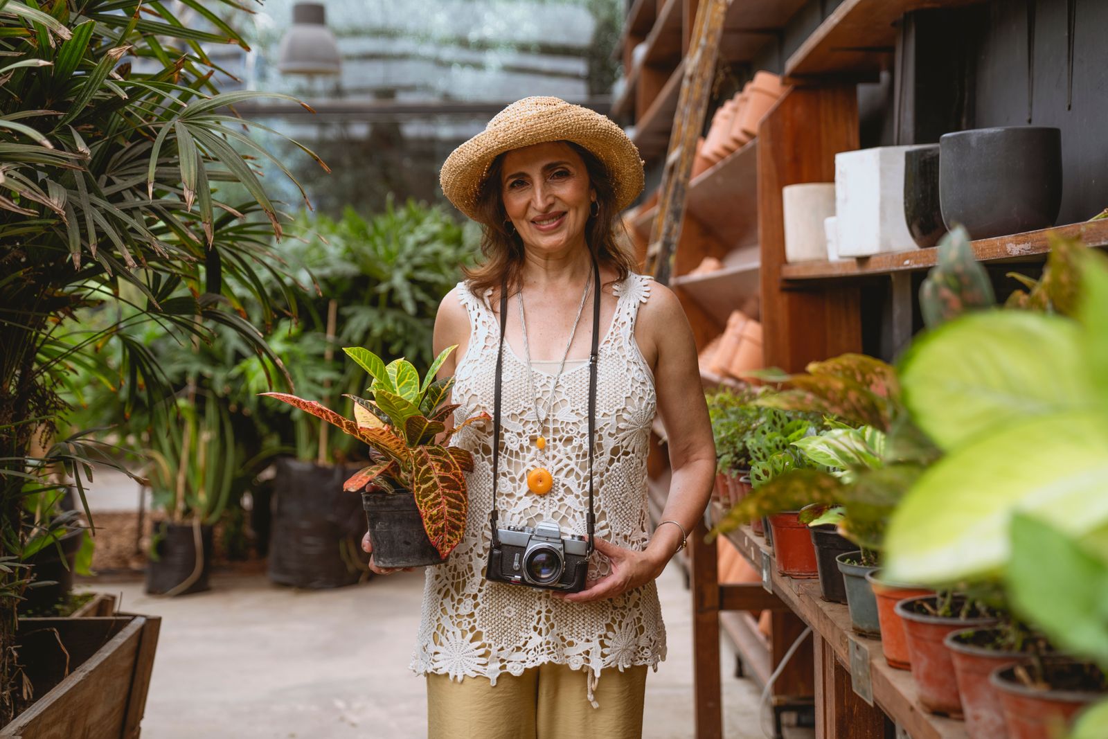 A gardener in a straw sun hat choosing a potted croton, terracotta and greenery on the shelves behind her.
