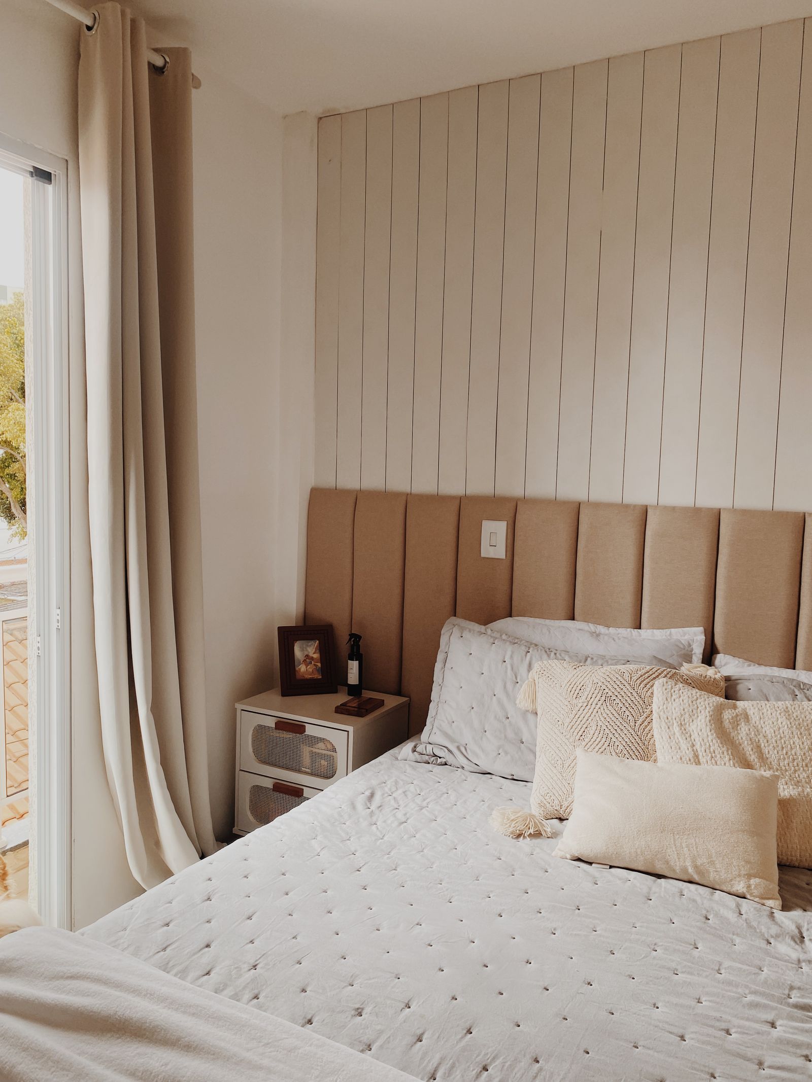 A bed made up in cream linens with a tan channel-tufted headboard, morning light through the balcony door.