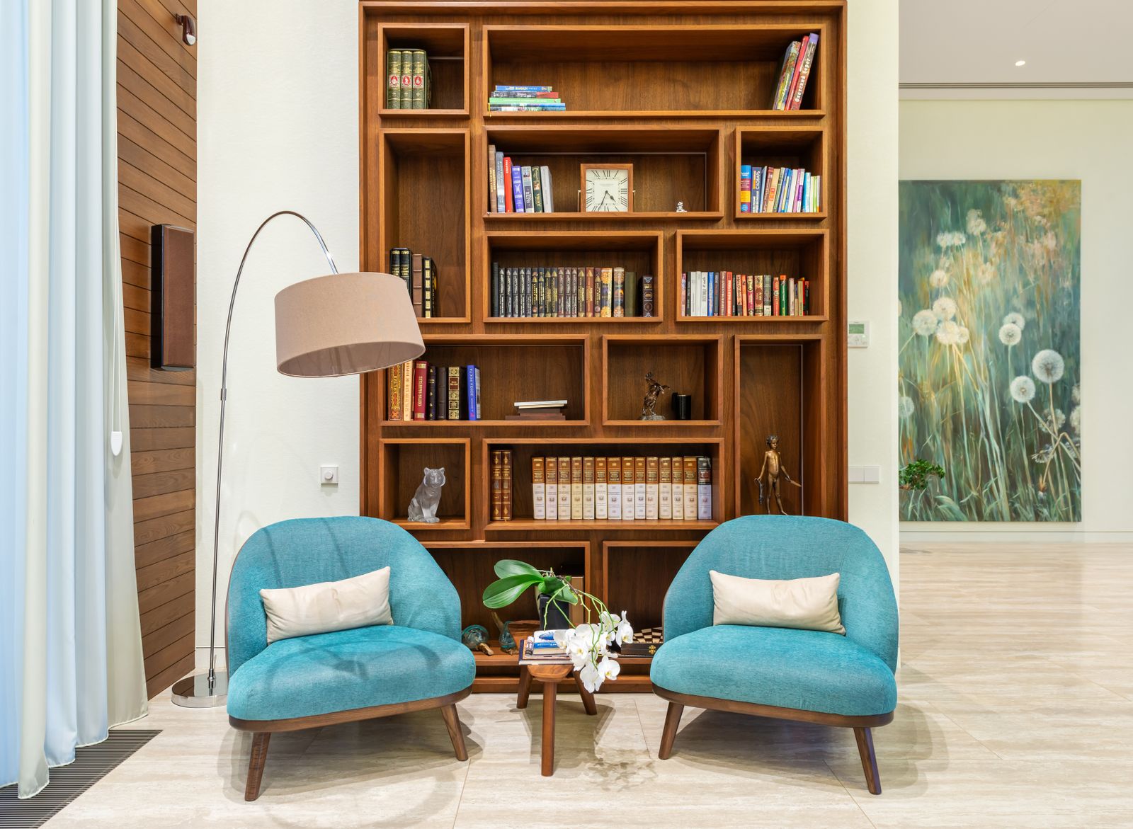 A tall walnut bookcase of open cubbies filled with books and a mantel clock, between two teal armchairs.