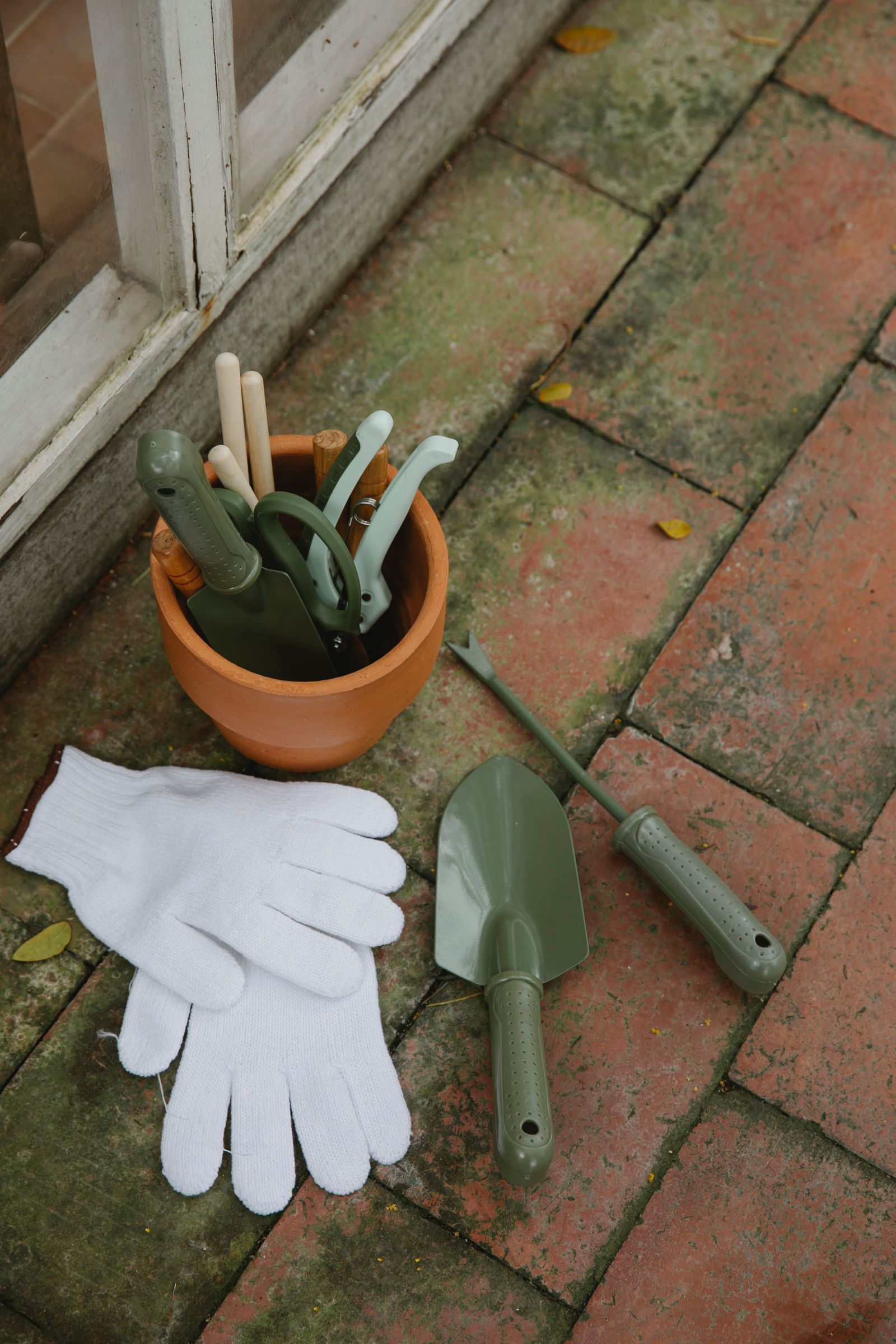 A terracotta pot of trowels and pruners beside a pair of knit gloves, set out on a mossy brick patio.